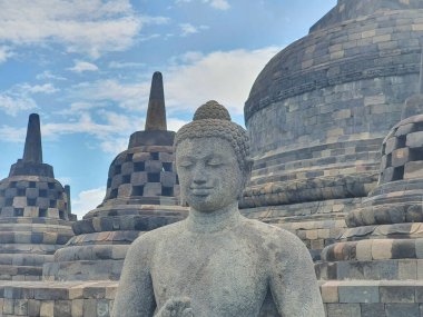 Yogyakarta 'daki Borobudur Tapınağı' nın Antik Taş Buda Heykeli Görkemli Tapınak 'ın önünde durgun duruyor. Mavi Gök' ün altında pofuduk Beyaz Bulutlar ve Kültürel Miras Mirası ile bir huzur sembolü..