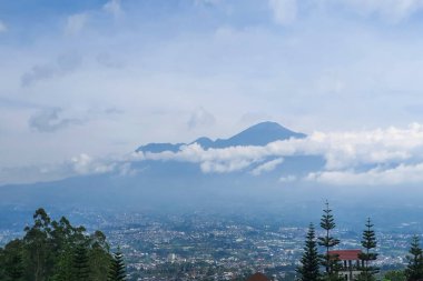 Majestic Mountain Views: Şehir manzarası bir tepeden ortaya çıktı, Bulutlar zarafetle sarmalandı, Gökyüzü altında sisli görünen uzak Skyline