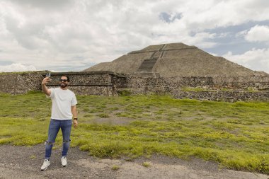 Genç adam arka planda fotokopi alanı olan Teotihuacan piramitleriyle akıllı telefonuyla selfie çekiyor.