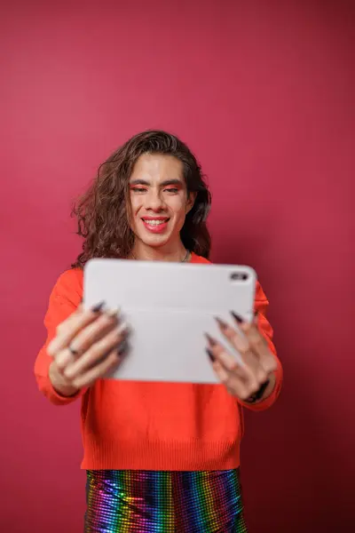 Kırmızı arka planda tablet kullanan ve gururlu kıyafetler giyen ikili olmayan kişi.