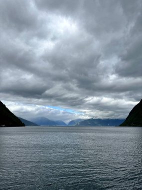 Sahne, arka planda dağlar olan bir su kütlesinin üzerinde bulutlu bir gökyüzü tasvir ediyor, bulutlar, su, gökyüzü ve dağlar gibi elementleri vurguluyor. Sognefjorden, Norveç