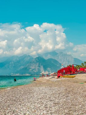 Türkiye 'nin Antalya kentindeki Akdeniz kıyılarının güzel manzarası. Açık bir günde, kırmızı güneş şemsiyeleri kayalık plaja karşı duruyor ve berrak mavi suya yansıyor. Destansı bulutlar