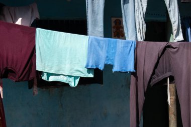 Gömlekler ve pantolonlar da dahil olmak üzere renkli bir kıyafet dizisi, mavi bir duvara asılı çamaşır ipine asılmış, güneş ışığı günlük hayatın basit bir kesimini gölgeliyor..