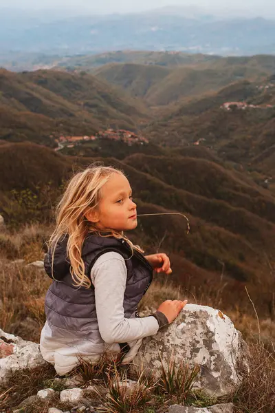 stock image a little blonde girl resting on a stone during a hike in the mountains against the backdrop of an autumn mountain landscape. Tuscany in autumn