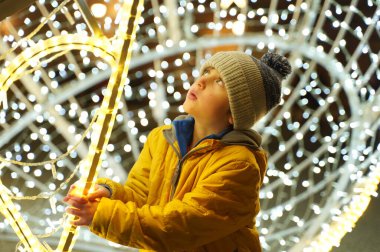 Mutlu Noeller ve mutlu yıllar. Sarı ceketli anaokulu öğrencisi elinde güzel bir Noel ışığı süslemesiyle meydanda duruyor. Gece şehrinde büyük bir Noel ağacı topu şeklinde.