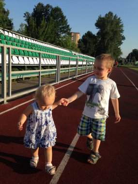 Küçük çocuklar stadyum koşu bandı boyunca koşuyorlar. Bir çocuk, kız kardeşinin koşmasına yardım ederken elini tutar.