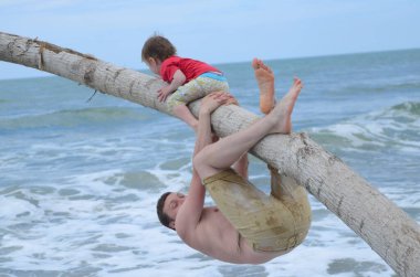 Above the waves, a father and son climb the trunk of a tree washed up on the shore clipart