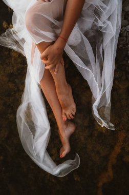 Kadının çıplak bacakları ince beyaz tül kumaşa sarılıp doğal bir taş yüzeyde dinleniyor. Huzurlu kompozisyon sükuneti ve zarafeti vurguluyor.