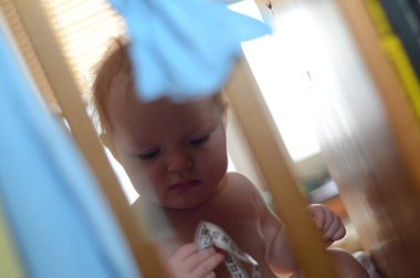 Bebek beşikte oturuyor, odaklanmış ve meraklı, mezurayla oynuyor. Yumuşak ışık, çocuğun konsantrasyonunu vurgular. Sahne bir çocukluk keşfi anını yakalıyor.