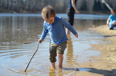Küçük çocuk göl kıyısında bir sopayla sığ suları zevkle keşfeder. Meraklı bir ifade, kaygısız bir eğlence, açık hava macerası sahilde güneşli bir günde