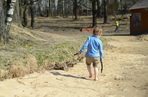 Mavi gömlek ve şort giyen genç bir çocuk kumlu bir yolda çıplak ayakla yürüyor, her iki elinde sopalarla. Arka planda ağaçlı bir park görünüyor.