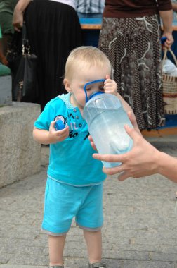 Turkuaz kıyafetli çocuk dışarıdaki büyük bir su şişesinden su içiyor. Asfaltlı bir alanda duran çocuk, yetişkin yardımı ile susuzluğunu gidermeye odaklanmış görünüyor.