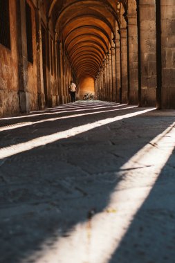 Kemerli taştan bir koridorda yürüyen tanınmayan biri yerdeki gölgelerle güçlü bir zıtlık yaratıyor. Uzakta iki bisiklet görülür ve görsel ilgi eklenir