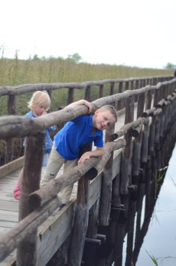 Çocuklar su üzerinde ahşap bir tahta köprüyü eğlenceli bir şekilde keşfediyorlar. Bir çocuk merakla ve heyecanla tırabzana yaslanırken, diğeri onu yakından takip ediyor.