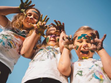 Çocuklar sevinçle renkli boyalı ellerini ve yüzlerini gösteriyor, heyecan ve yaratıcılıklarını ifade ediyorlar. Benekli gömlekler giyiyorlar, dışarıda eğlenceli ve sanatsal bir oyun oynuyorlar.