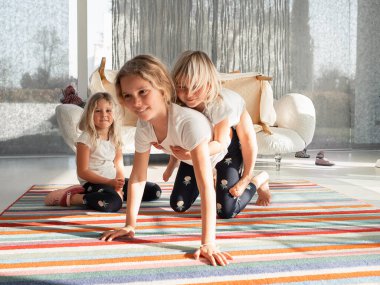 Üç kız kardeş parlak çizgili bir halıda şakacı hareketler yapıyorlar. Üçüncüsü yakınlarda oturup gülümserken iki çocuk sırtında oyun oynuyor.