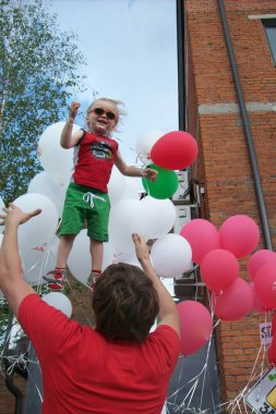 Renkli kıyafetler giyen ve güneş gözlüğü takan genç çocuk elinde renkli balonlar tutarken neşeyle havaya savruluyor. Aşağıdaki baba çocuğu destekliyor, hayat dolu bir çevreyle çevrili.