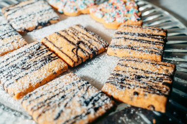 Çikolata serpiştirilmiş, toz şeker serpiştirilmiş, gümüş tepsiye konmuş kare kurabiyeler. Arkaplanda renkli serpiştirilmiş dekoratif kurabiyeler var.