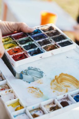 Canlı bir boya paletinin renklerini karıştırmak için fırça kullanan bir elin yakın çekimi, sanatsal bir ortamda boyaları karıştırma sürecini yakalıyor.