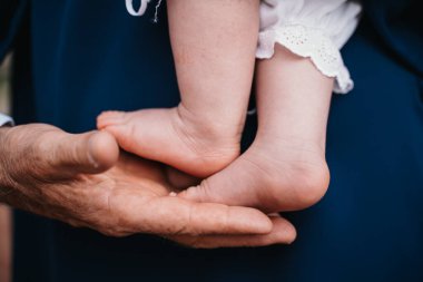 Yaşlı eller bebek ayaklarını nazikçe tutarak nesiller boyu bakım ve korumayı sembolize ediyor. Yaşlanmış cilt ve yumuşak, narin bebek ayakları arasındaki zıtlık sıcaklığı ve bağlantıyı yakalar.