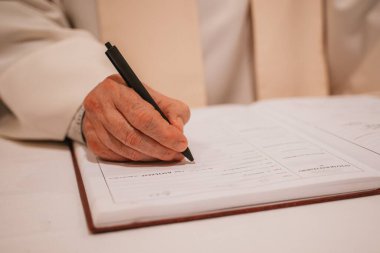 Kilisenin kayıt defterine kalem kullanarak vaftiz detaylarını yazan bir rahibin yakın çekimi. Sayfa vaftiz belgeleriyle ilgili bölümler içeriyor, bir ayin sürecini yansıtıyor.