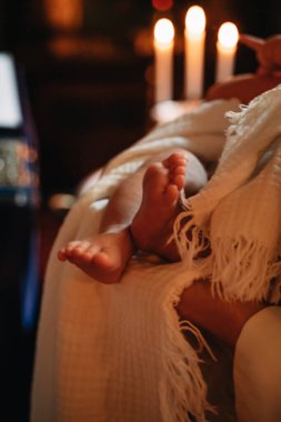 Bir kilise ayininde vaftiz töreni sırasında beyaz beze sarılmış bir bebek. Arka plandaki mum ışığı kutsal ana sıcak ve huzurlu bir atmosfer ekler..
