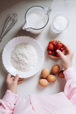 Pişirme malzemeleri un, şeker, süt, yumurta ve taze çilek de dahil olmak üzere mutfak tezgahında güzelce düzenlenir. Ellerinde bir tabak un ve bir tabak çilek var..
