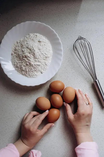 Eller bir kase un ve mutfak tezgahında çırpılmış bir çırpıcının yanına yumurta yerleştiriyor. Bu da pişirme hazırlığını gösteriyor. Sahne ev yemeği aktivitesi olduğunu gösteriyor.
