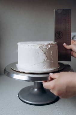 Elleri metal bir cetvel tutarak pikabın üzerindeki beyaz kremalı kekin boyunu ölçüyor. Görüntü pasta dekorasyonu ve pişirme tekniklerindeki hassasiyete odaklanıyor.