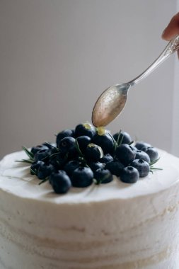 El, beyaz kremalı pastaya taze yaban mersini ve limon lezzeti ekleyerek görsel olarak çekici bir tatlı yaratıyor. Canlı renkler ve dokular pastanın sunumunu güçlendirir