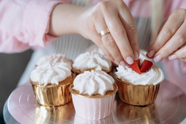 Eller, krem şantili keklerin üzerine taze çilek dilimleri diziyor. Görüntü, görsel olarak çekici bir sunum için tatlıları dekore etme sürecini yakalar.