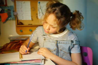Masasında bir deftere ödev yazan odaklanmış, saçları örgülü, çizgili gömleği ve kot tulumu olan, renkli kırtasiye ve kitaplarla çevrili genç bir kız.