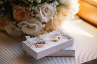 Altın alyanslar zarif bir şekilde beyaz bir hediye kutusunda sergileniyor bir fiyonkla süslenmiş, arkaplanda kaymak gibi güller sergileniyor. Aşk ve bağlılığın güzel bir sembolü, düğün teması için mükemmel.