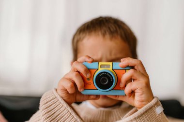Genç çocuk canlı bir oyuncak kamera tutuyor, fotoğraf çekiyormuş gibi yapıyor. Kameranın parlak renkleri ve çocuk dostu tasarımı onu yaratıcı oyun için ideal kılıyor.