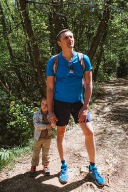 Bir babanın ve oğlunun birlikte ormanı keşfetmesinin unutulmaz anı. Çocuk babasının elini tutuyor, meraklı ve meraklı görünüyor, baba ise yukarıya bakıyor, muhtemelen çevreyi gözlemliyor. Bir macera ve bağlılık duygusu sergiliyorlar.