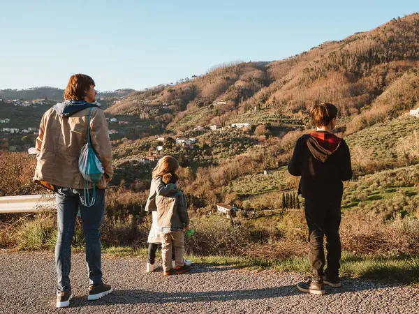 Aile, dağ manzarasının nefes kesici görüntüsünü alır. Baba ve çocuklar bir arada, güneşli bir günde doğanın dingin güzelliğinin tadını çıkarıyorlar. Deniz mavisi sırt çantası ve günlük giysiler bir macera ve rahatlama günü olduğunu gösteriyor.
