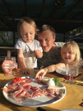 Mutlu aile, açık havada gün ışığında piknik yaparken bir tabak İtalyan pastırması yiyor. İki oğul ve baba tabağındaki et ve peynir çeşitliliğine heyecanla bakıyorlar. Sahne, yemek ve neşe dolu bir anı yakalıyor. 