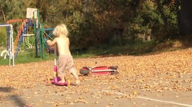 Çıplak ayaklı ve meraklı bir çocuk, iki pembe scooterlı bir sonbahar parkını keşfediyor. Düşen yapraklar ve ağaçlarla çevrili sahne, neşeli keşif ve masumiyetin özünü yakalıyor.