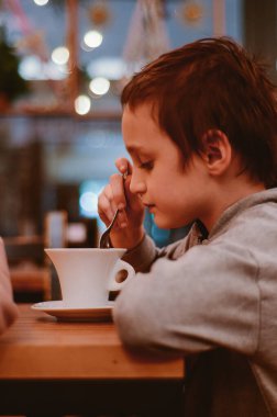 Derin düşüncelere dalmış genç bir çocuk sıcacık bir kafe ortamında sıcak bir içecek karıştırıyor. Yumuşak ışıklandırma ve samimi ortam sıcak ve davetkar bir atmosfer yaratır.