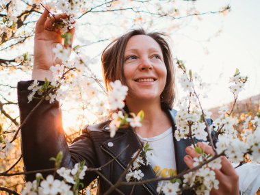 Güneşin doğuşunda kiraz çiçekleriyle çevrili gülümseyen kadın huzurlu havanın tadını çıkarıyor. Deri bir ceket giyiyor, ılık ışıkta güneşlenirken dallara hafifçe dokunuyor..