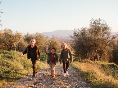 Bir grup çocuk, zeytin ağaçlarıyla çevrili manzaralı bir dağ yolunda mutlu bir yürüyüş yapıyorlar. Çocuklar mutluluğu ve macerayı ifade eder, doğada mükemmel bir sonbahar gününü yakalarlar..