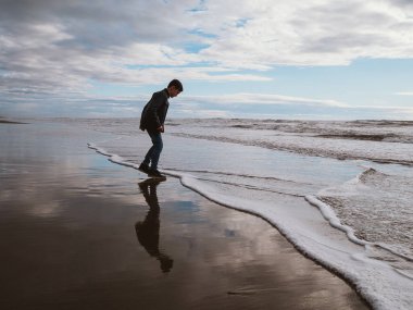 Genç bir çocuk bulutlu bir sahil gününde yumuşak okyanus dalgalarına adım atıyor. Gökyüzünün ve kıyı şeridinin yansıması rahatlatıcı kavramlar için mükemmel sakin ve huzurlu bir manzara yaratır.