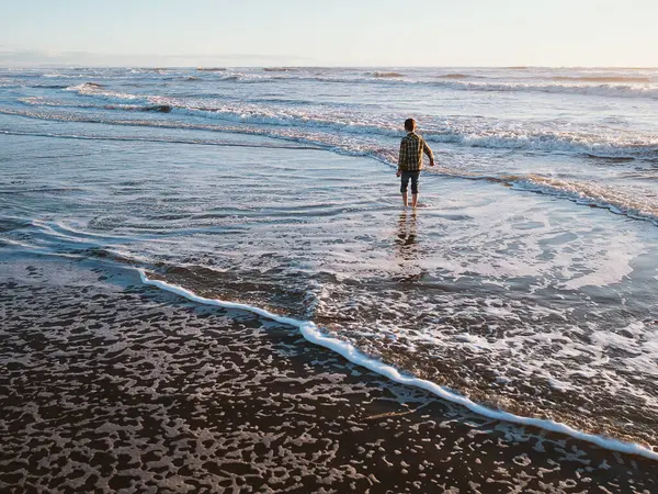Genç bir çocuk güneşli bir sahil gününde yumuşak deniz dalgalarına adım atıyor. Gökyüzünün ve kıyı şeridinin yansıması rahatlatıcı kavramlar için mükemmel sakin ve huzurlu bir manzara yaratır.