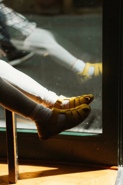 Parlak sarı sandaletler ve beyaz taytlar giyen bir çocuk bir pencerenin yanında rahatlar. Güneş ışığı neşeli gölgeler oluşturur, sandaletleri renklendirir ve sıcak bir atmosfer yaratır.