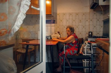Mutfak masasında tek başına oturan yaşlı bir kadın, ev eşyalarıyla çevrili. Onun düşünceli ifadesi günlük hayatta bir yalnızlık hissi taşır. Sıcak, sıcak bir atmosfer.