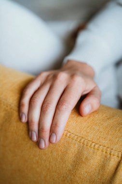 Bir insanın elini, rahat sarı bir kanepenin kolunda nazikçe dinlendirirken yaklaş. Görüntü, kumaşın dokusunu vurgulayarak dingin ve gevşetilmiş ortama odaklanır..