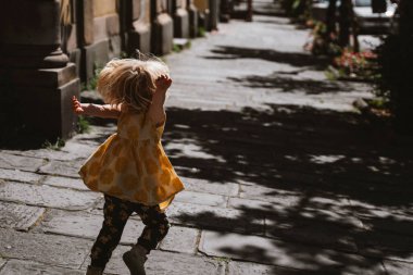 Küçük çocuk neşeyle güneşli bir caddede koşuyor, sarı bir elbise giyiyor, etrafı gölgeler ve bir şehrin ışığıyla çevrili. Sahne, özgürlük ve oyun hissini yakalar..