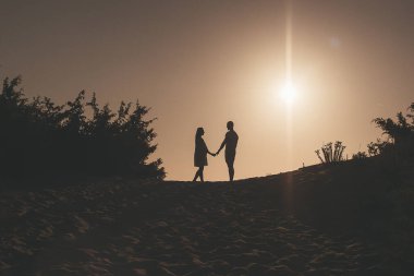 Güneş arka planda batarken kum tepeciğinde el ele tutuşan bir çiftin silueti. Sahne, onların sıcak etkileşimleriyle vurgulanan romantizm ve bağlantı duygularını çağrıştırıyor.