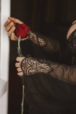 Siyah dantel kollu narin eller pencerenin yanında canlı kırmızı bir gül tutuyor. Karmaşık dantel detayı zarafet ekler, kırmızı gül ise tutkuyu ve aşkı simgeler..