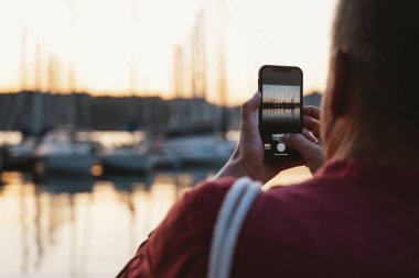 Şahıs limanda sakin bir günbatımını fotoğraflamak için akıllı telefon kullanıyor, tekneleri ve sakin suları vurguluyor. Sahne, doğanın değerini arttıran teknolojinin altını çiziyor..